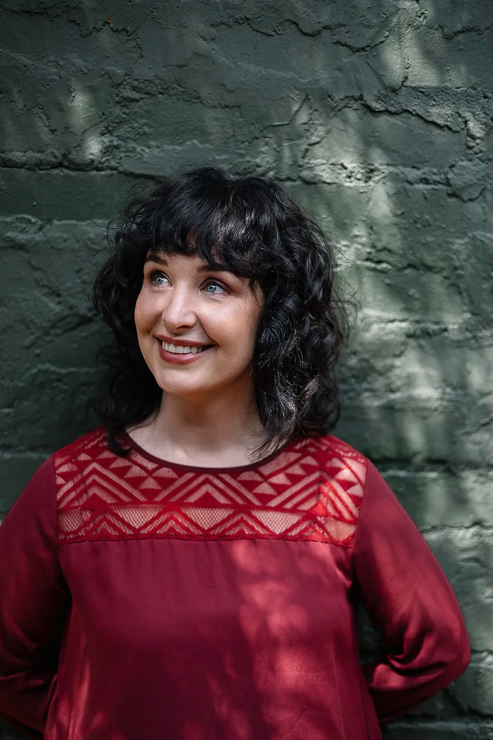 Elri Kuhn smiling and looking up, in front of a green painted brick wall